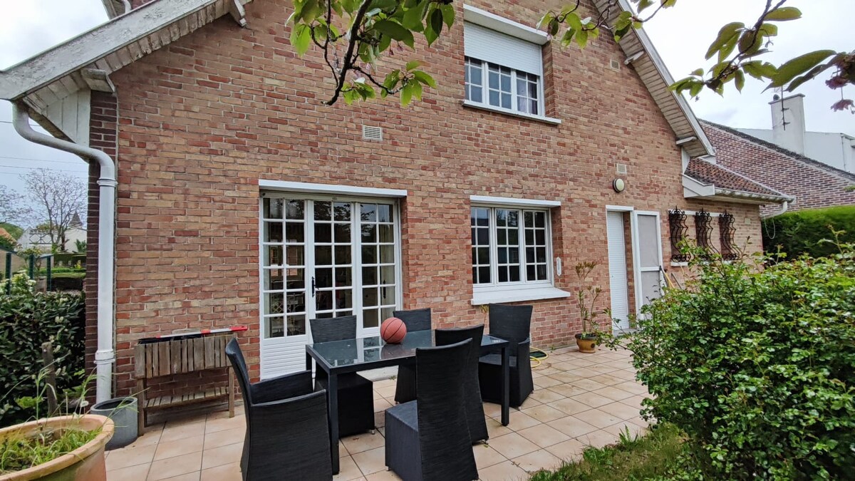 A brick exterior of the house is featured, showcasing large glass doors leading to a terrace. Outdoor furniture includes a glass dining table surrounded by dark wicker chairs. Lush greenery and potted plants enhance the serene garden space.