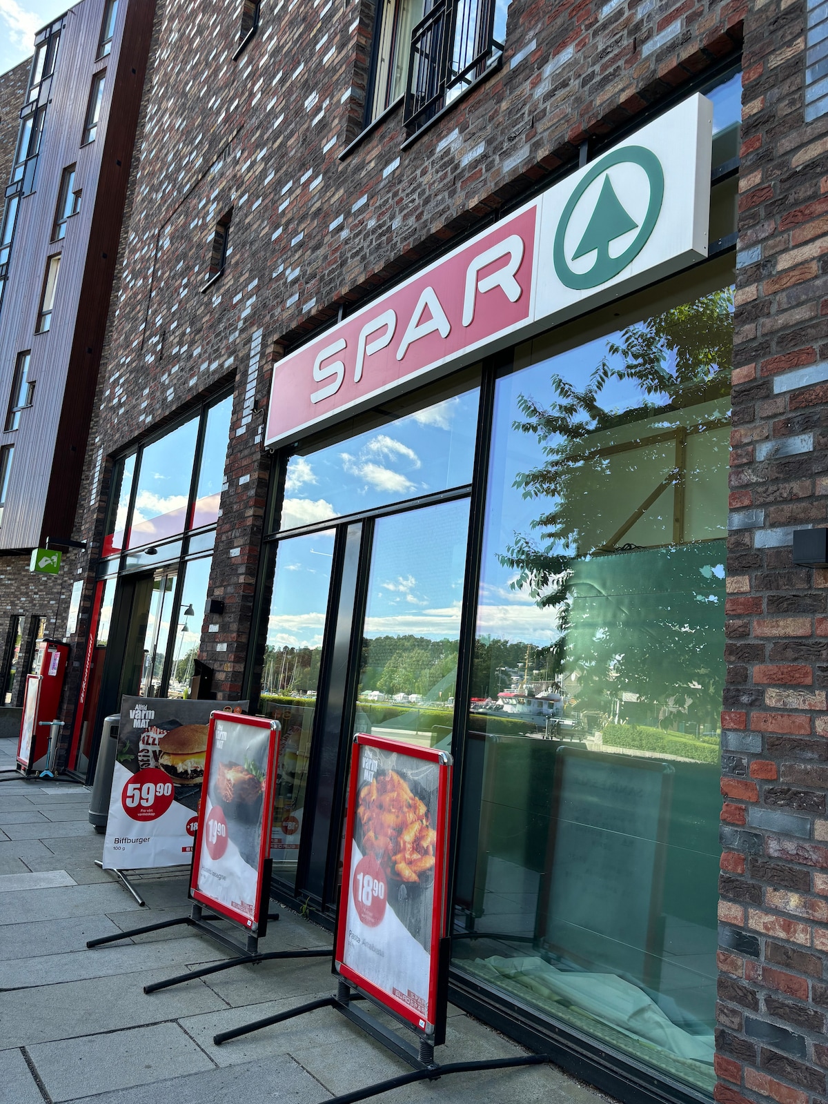 The entrance of a Spar grocery store is visible, featuring large glass windows that reflect the surroundings. Two sandwich boards display menu items outside, and the store logo is prominently displayed above the entrance. The area is paved, and nearby trees can be seen.