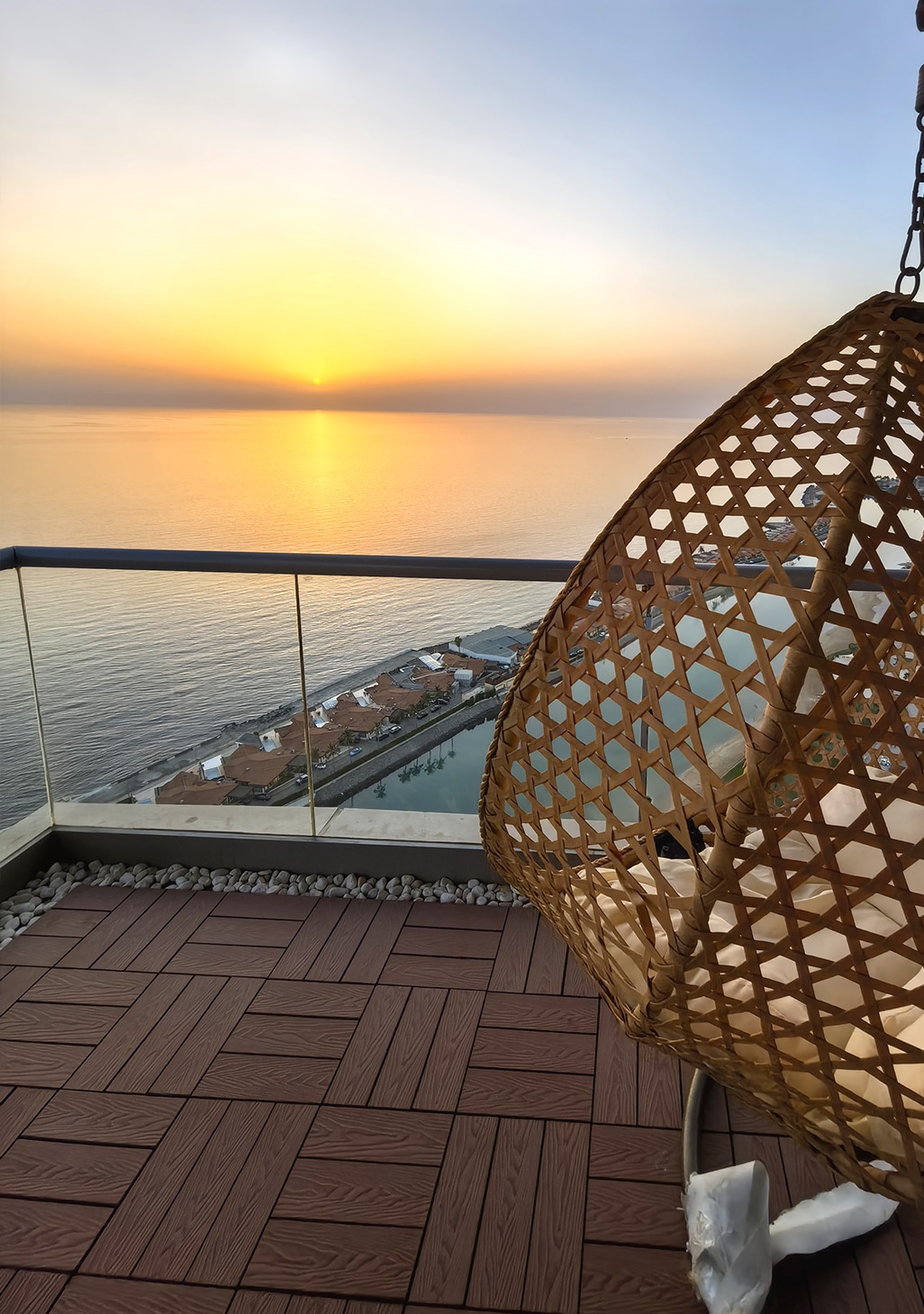 A hanging wicker chair is positioned on a balcony with a panoramic view of the sea at sunset. The golden hues of the setting sun reflect on the calm water, while the shoreline is visible below, enhancing the serene atmosphere.