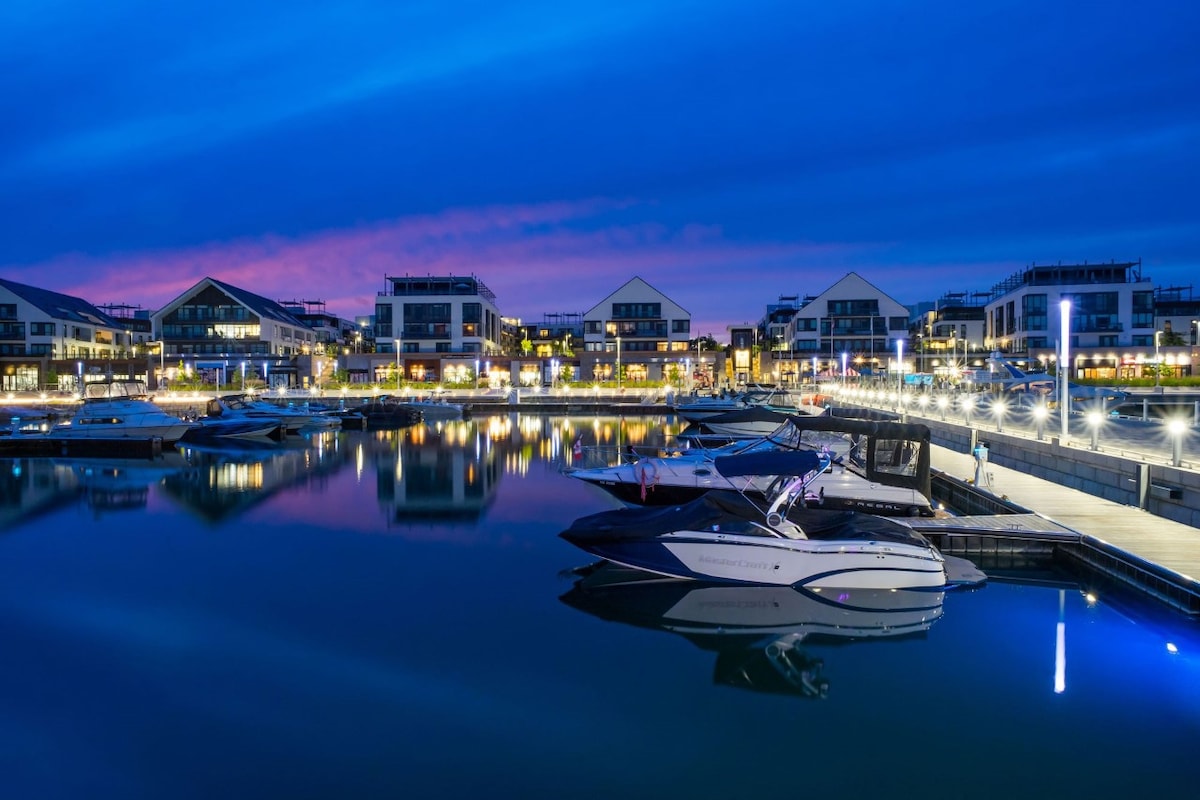 A serene marina at dusk is depicted with boats gently anchored along the pier. Soft lights reflect off the calm water, highlighting the modern buildings in the background against the twilight sky. The scene captures a tranquil atmosphere, ideal for relaxation.