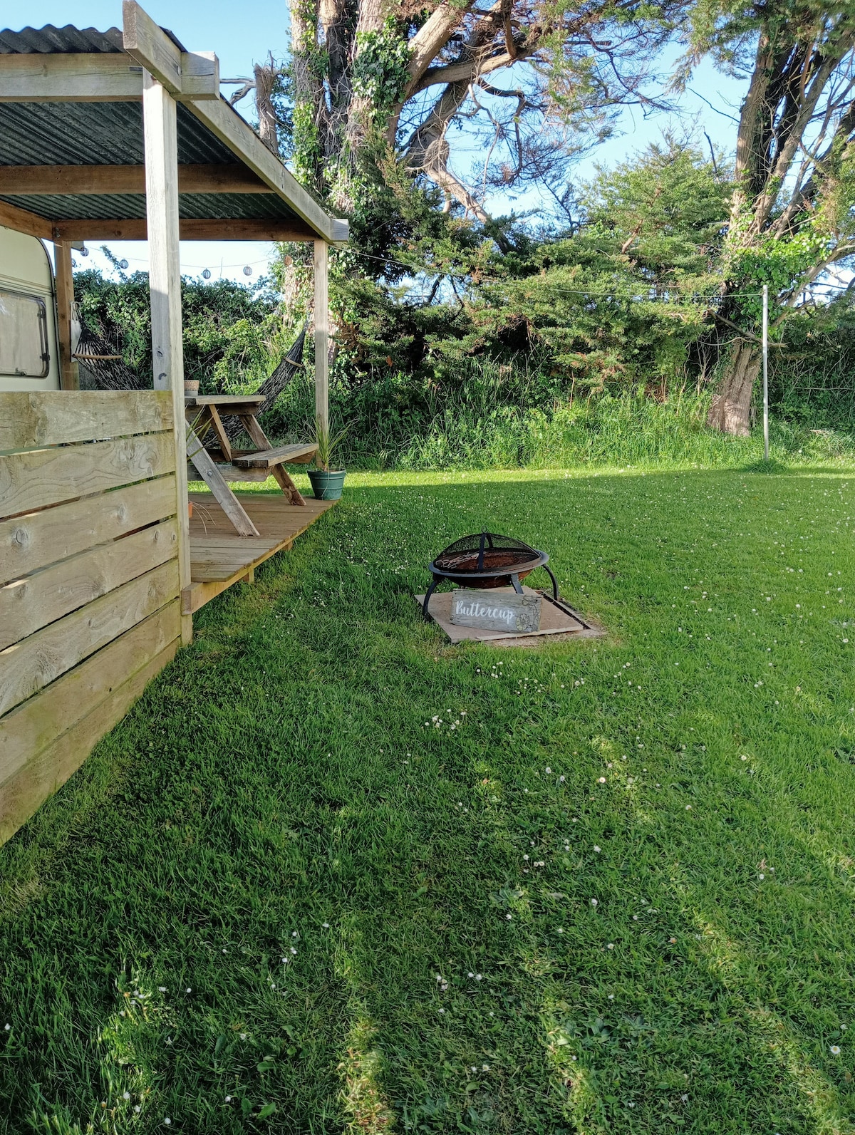 A grassy outdoor area is featured, showcasing a fire pit with a wooden seating area nearby. The scene captures the rustic charm of the caravan setting, framed by trees in the background, creating a serene environment for outdoor relaxation.