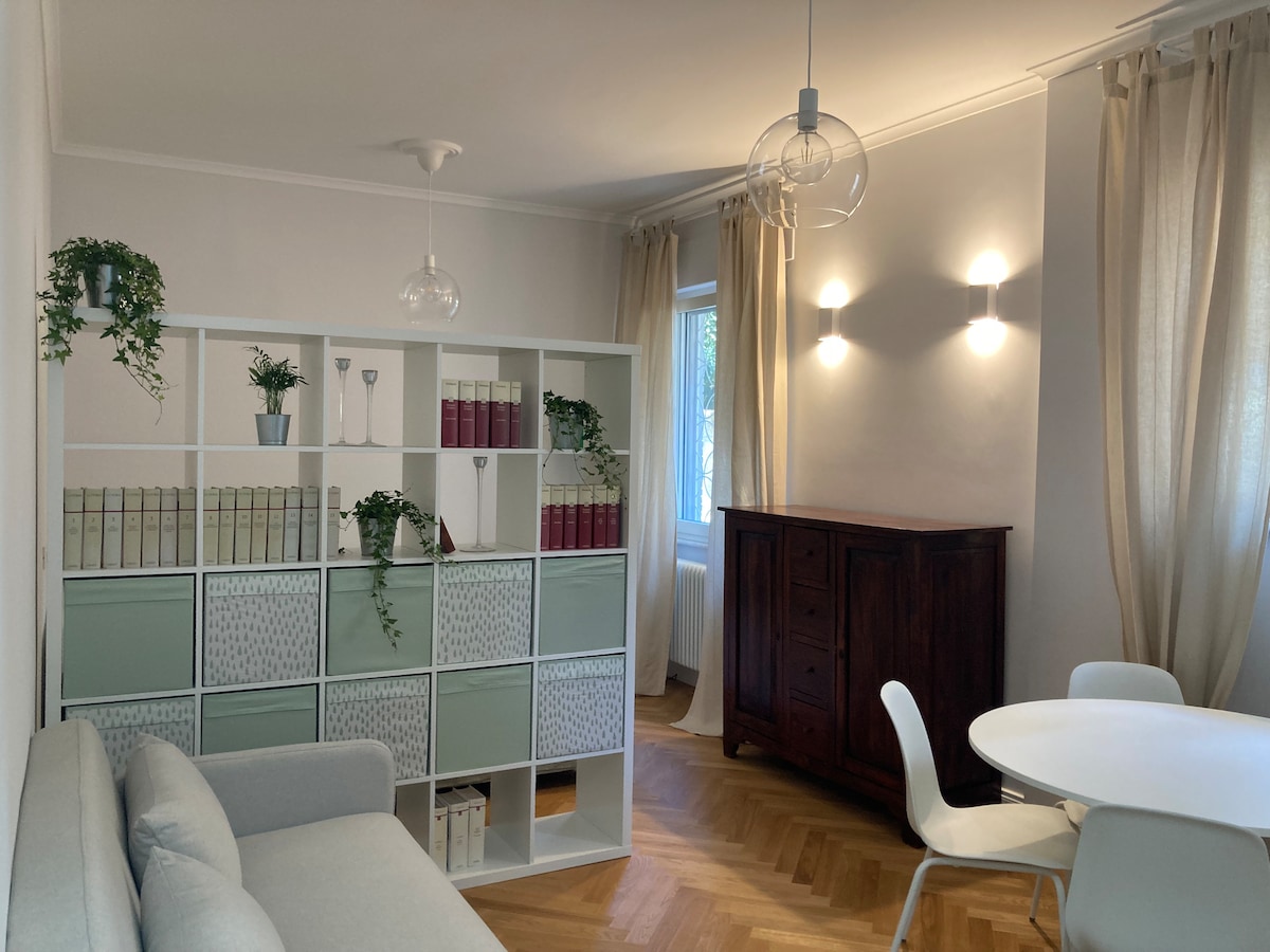 The living space features a light-colored sofa alongside a bookshelf with decorative greenery and neatly arranged books. A round dining table sits near a wooden cabinet, while soft lighting emanates from modern wall sconces, creating a calm ambiance. Natural light enters through the window dressed with sheer curtains.