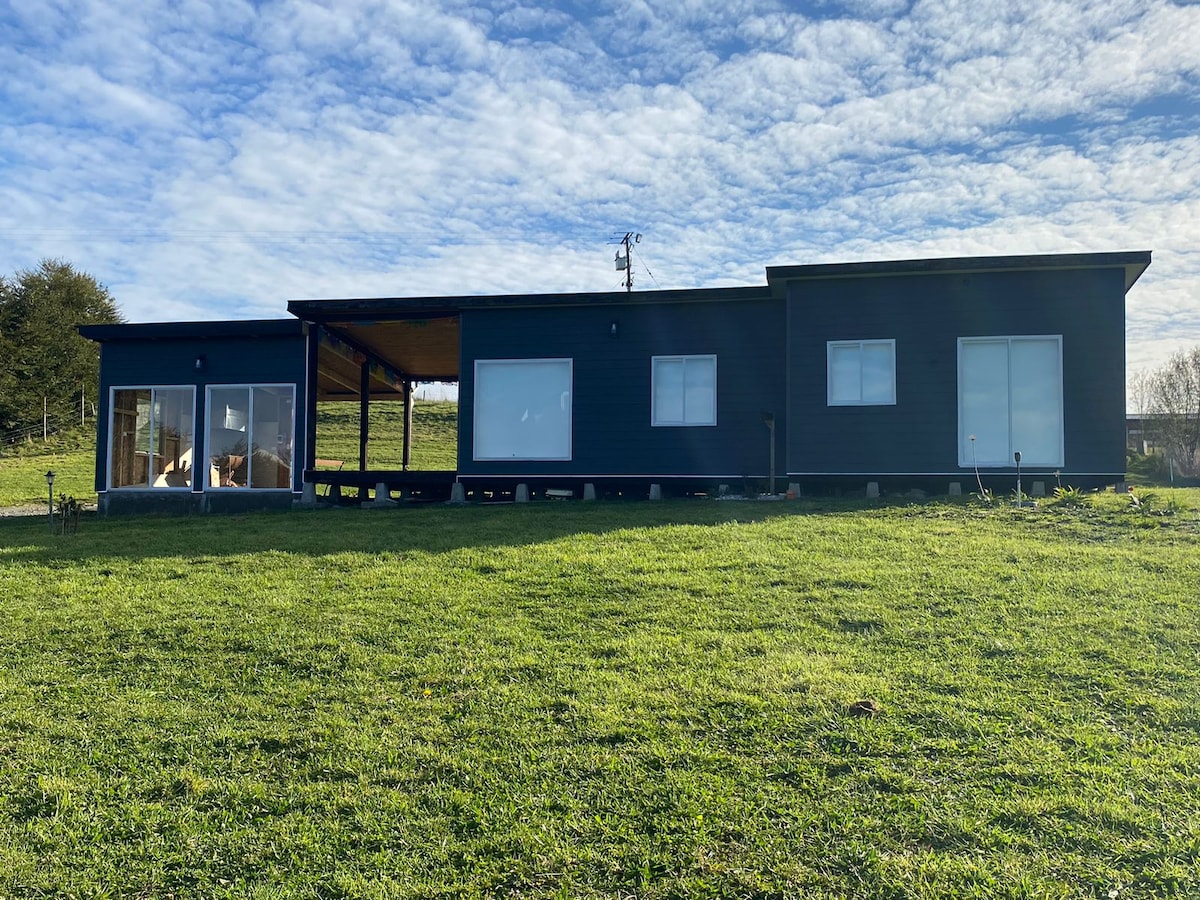 The image displays a modern single-story house set against a backdrop of rolling green fields. Large windows provide ample natural light, while a covered porch area invites outdoor relaxation. The structure features a sleek dark exterior, contrasting with the bright sky above.