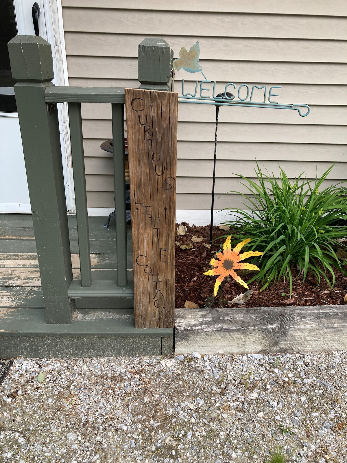 A wooden welcome sign is displayed on the porch railing, inscribed with 'Curious' and 'Haven.' A metal stake featuring a welcoming phrase and a colorful leaf decoration is positioned nearby. Lush greenery and mulch enhance the outdoor environment, creating an inviting entrance.