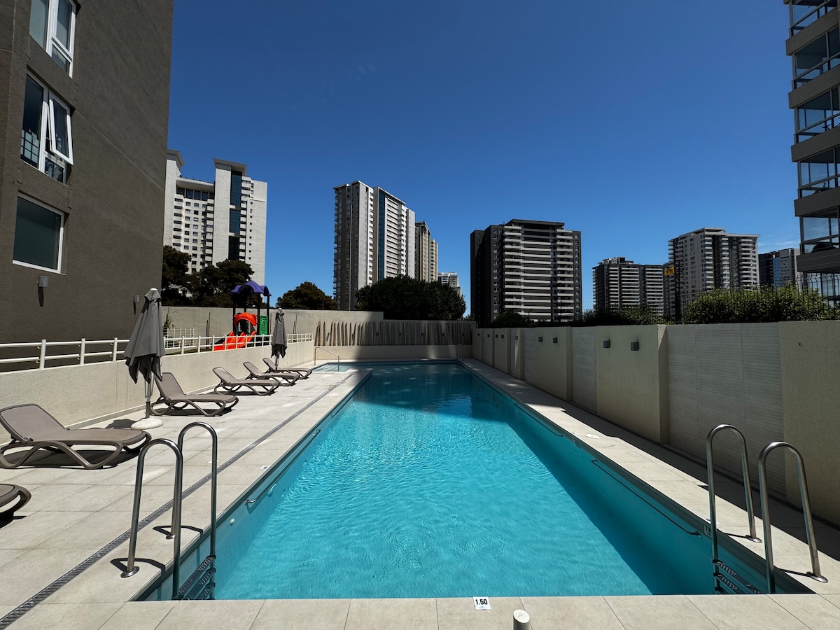 An outdoor heated pool features clear blue water, surrounded by a tiled deck equipped with several loungers. Tall buildings rise in the background, providing a modern urban backdrop against a bright blue sky.