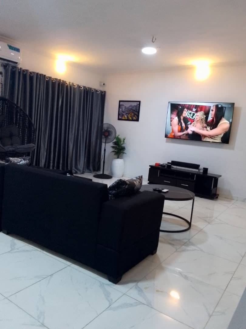 A modern living area features a dark sofa facing a large wall-mounted television. Soft lighting is provided by overhead fixtures and wall-mounted lamps. Dark curtains frame a window, while a white potted plant and a black coffee table complete the space, enhancing its welcoming appeal.