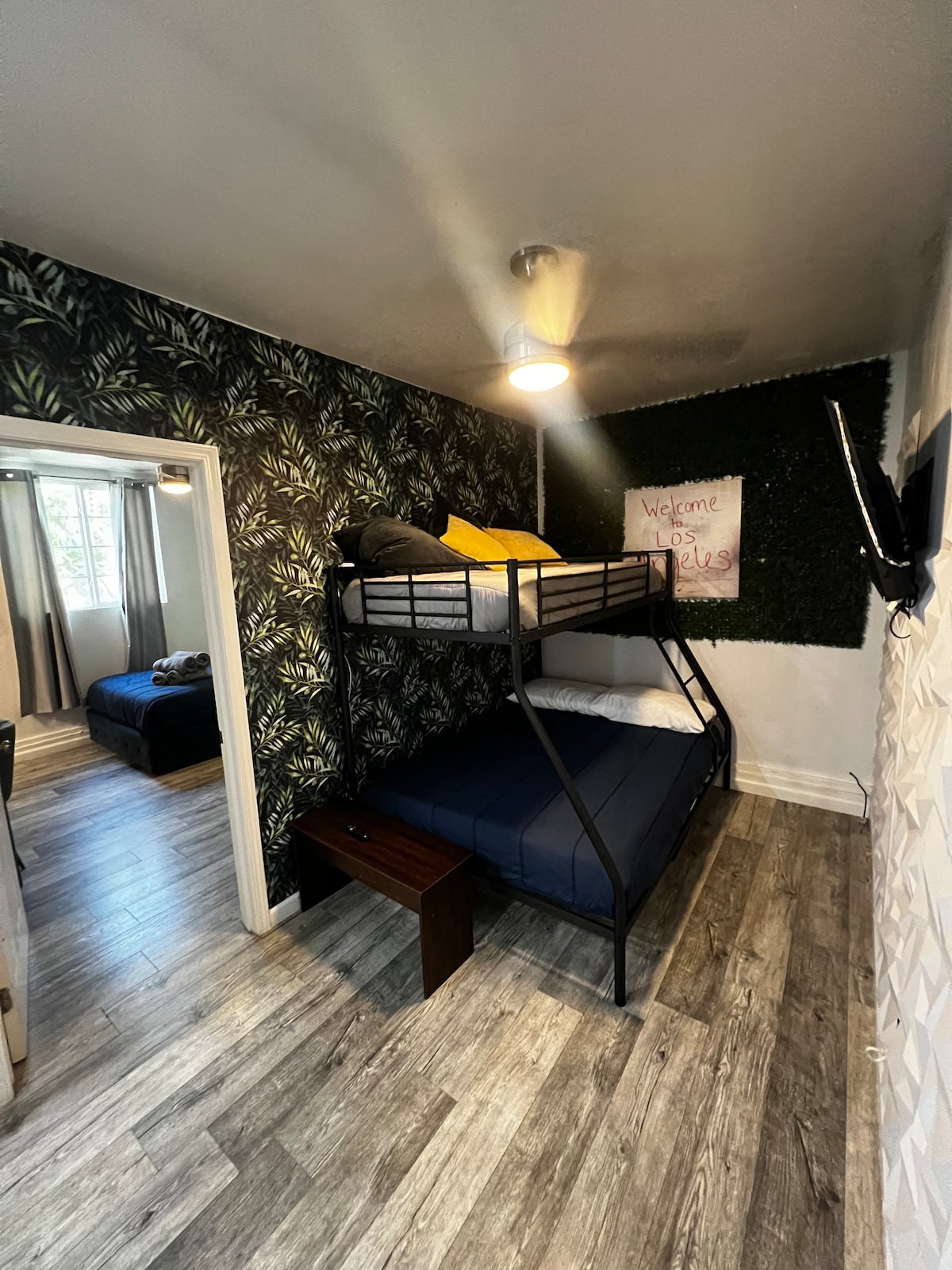 A functional sleeping area features a sturdy bunk bed with a dark blue mattress on the bottom and a black frame. A small wooden bench is positioned beside the bed. The walls are adorned with a leafy wallpaper, and natural light enters through a nearby window.