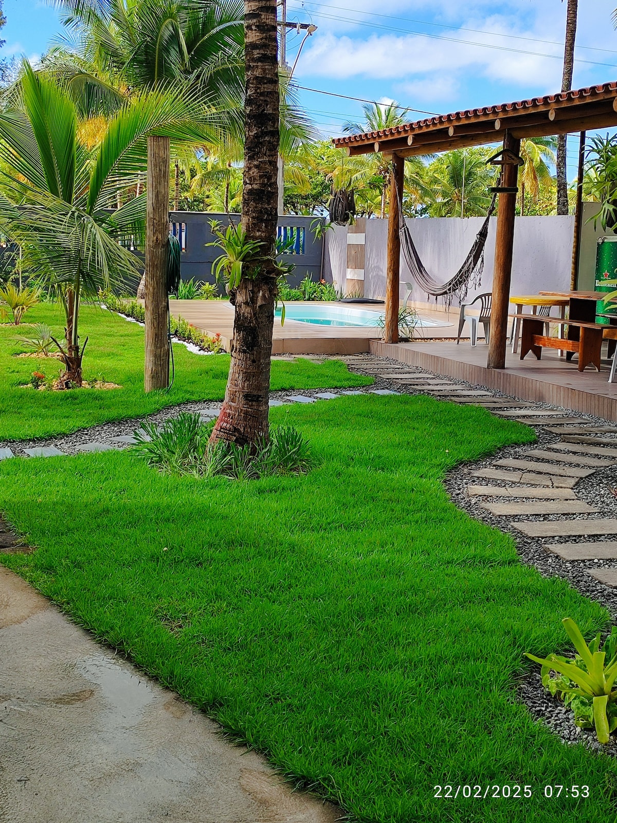 A well-maintained garden features lush green grass and palm trees, leading to a wooden patio area. A hammock is suspended near the patio, and a pool can be seen in the background. The pathway is lined with neatly arranged gravel stones, creating a welcoming outdoor space.