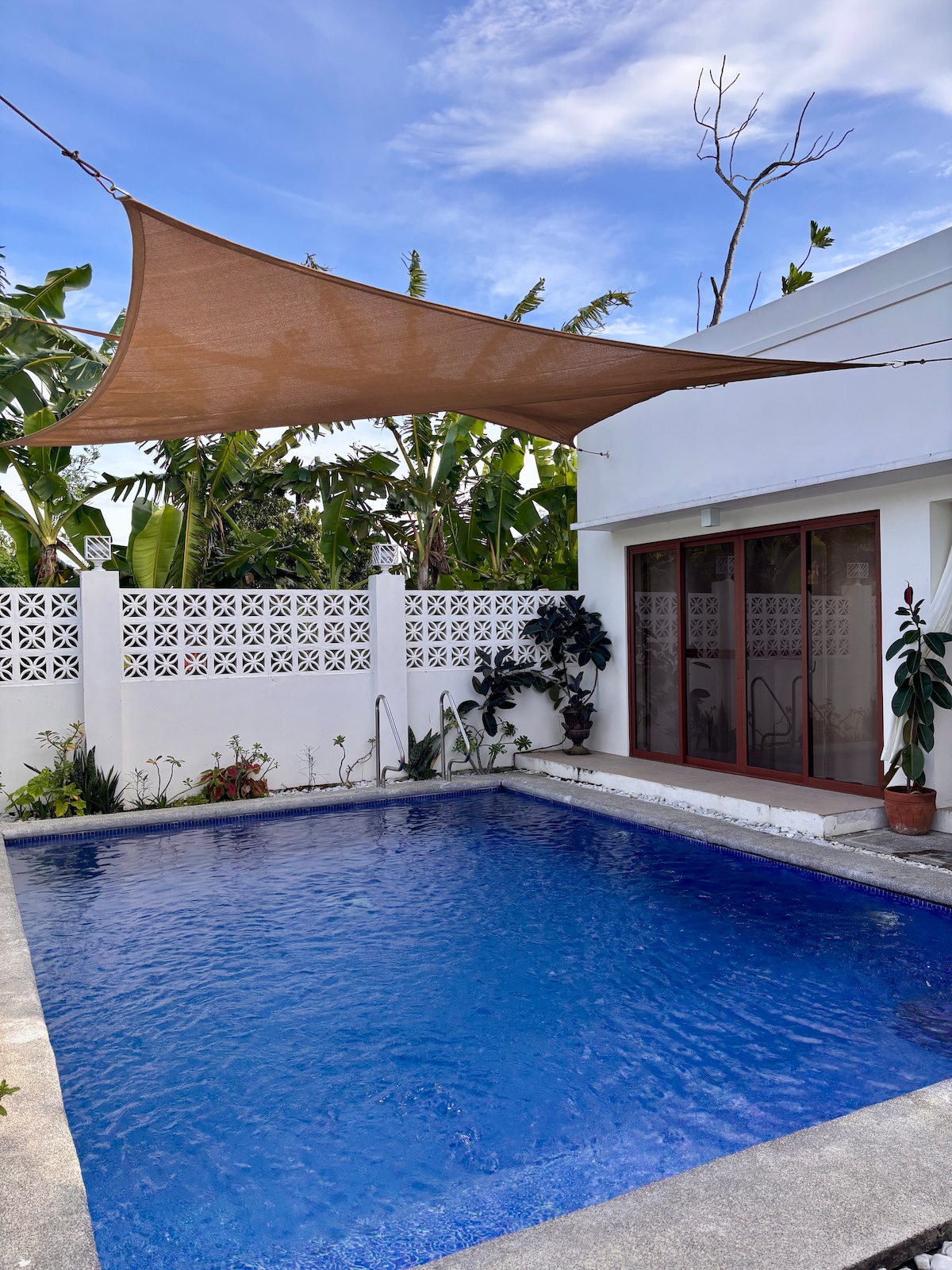 A serene swimming pool is surrounded by lush greenery and adorned with a sunshade overhead. The pool features clear blue water and is bordered by a smooth concrete deck, providing a calming outdoor space for relaxation.