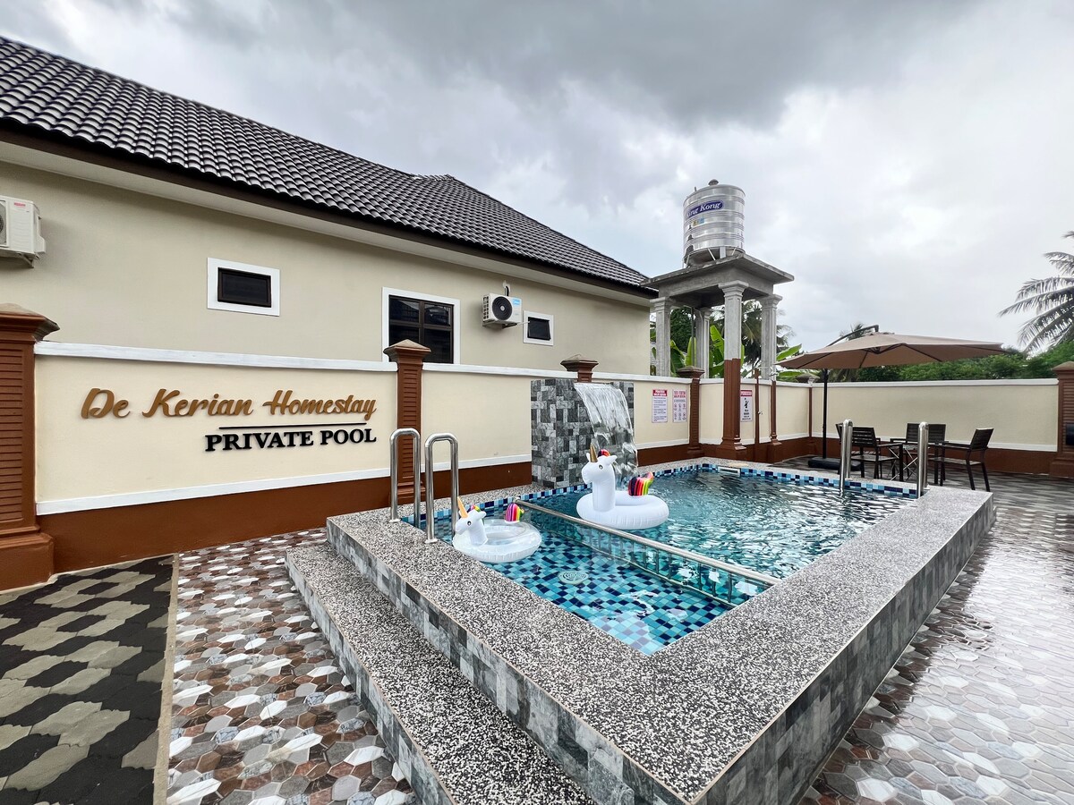 A private pool is featured, surrounded by a patio with decorative tiles. A small seating area is visible alongside the pool, and two floatation devices are in the water. The building has a neutral exterior with a sign indicating "De Kerian Homestay".