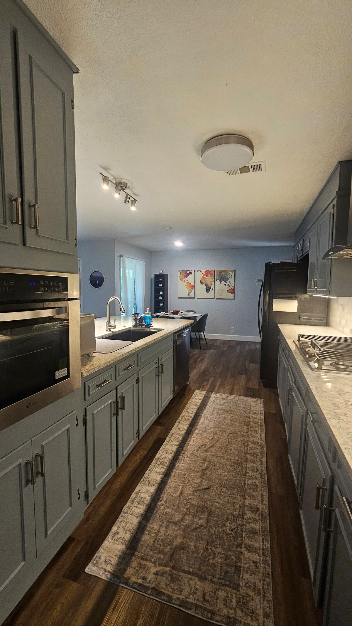 A well-equipped kitchen is presented with grey cabinetry and a long countertop featuring modern appliances. Light streams in from a nearby window, illuminating the space. A dining area is visible in the background, adorned with artwork that adds a touch of color.