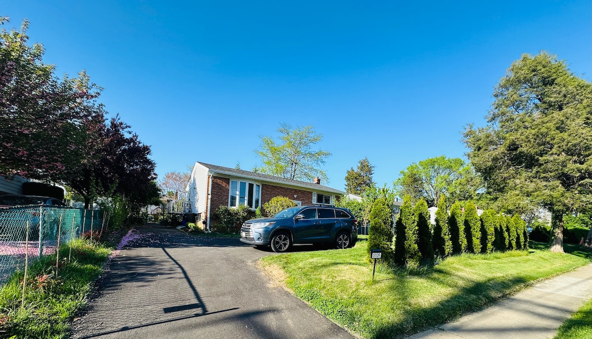 A single-story home is set in a well-maintained yard, surrounded by greenery and mulch beds. A paved driveway leads to the front of the house, and a dark-colored vehicle is parked to the side. Bright blue skies add to the welcoming exterior.