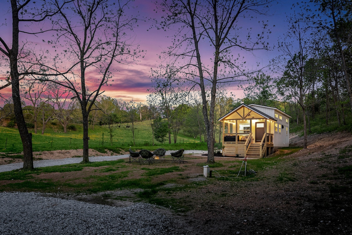The exterior of a charming tiny house is showcased at dusk, with glowing windows and steps leading to a large covered porch. A fire pit and seating area are visible in the foreground, surrounded by trees and open green fields, all under a colorful sunset sky.