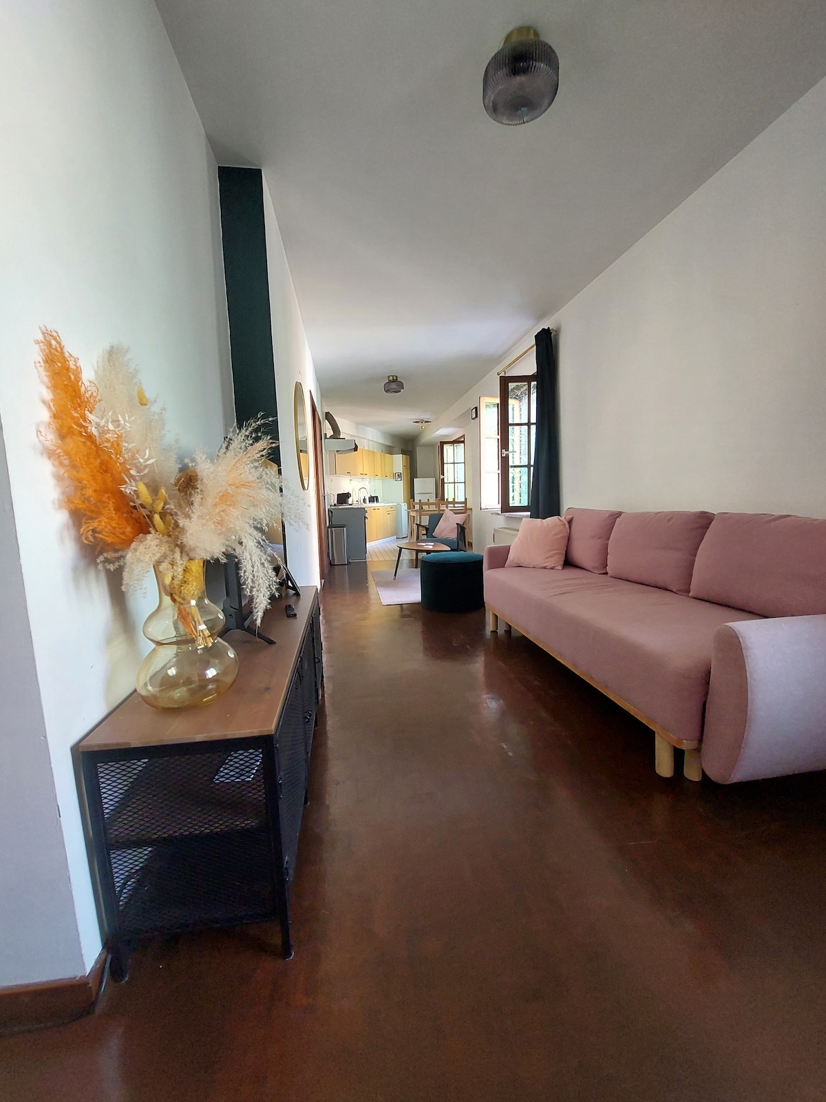 A long hallway displays a soft pink sofa and a decorative vase filled with dried plants. Light streams in through multiple windows, illuminating the open layout that leads to a well-equipped kitchen visible at the end.