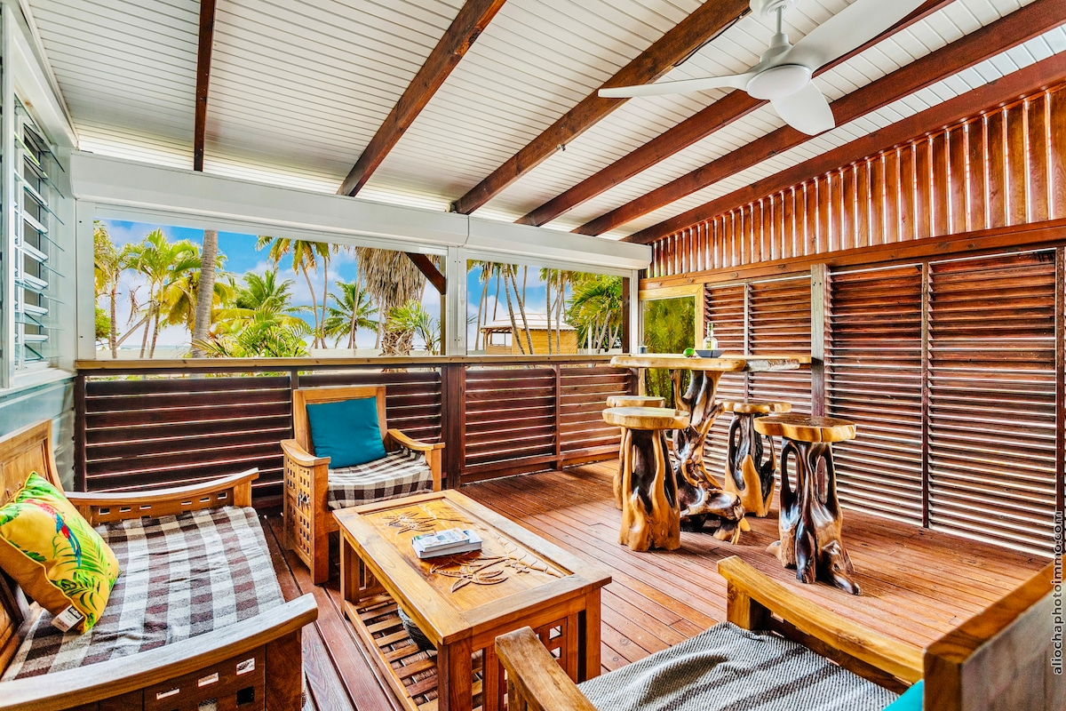 A spacious patio area is surrounded by wooden slatted walls, offering a view of palm trees and the beach beyond. Natural wood furniture, including tables and chairs, complements the warm tones of the deck. A ceiling fan is visible, enhancing comfort in this inviting space.