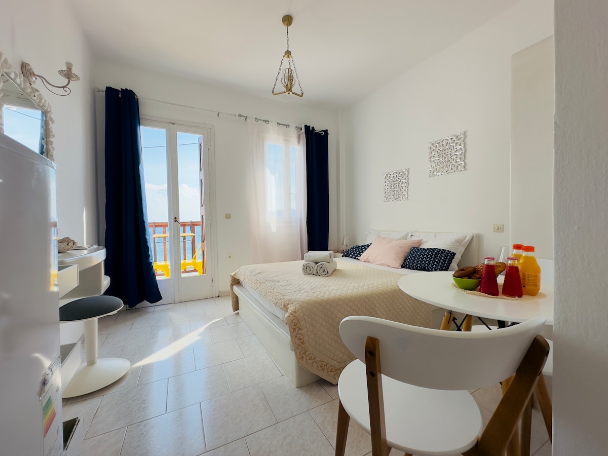 A bright sleeping area is featured, with a spacious bed dressed in neutral tones. A small round dining table with chairs is positioned nearby. Large windows allow natural light to illuminate the room, while blue curtains frame the view of the sea beyond the balcony.