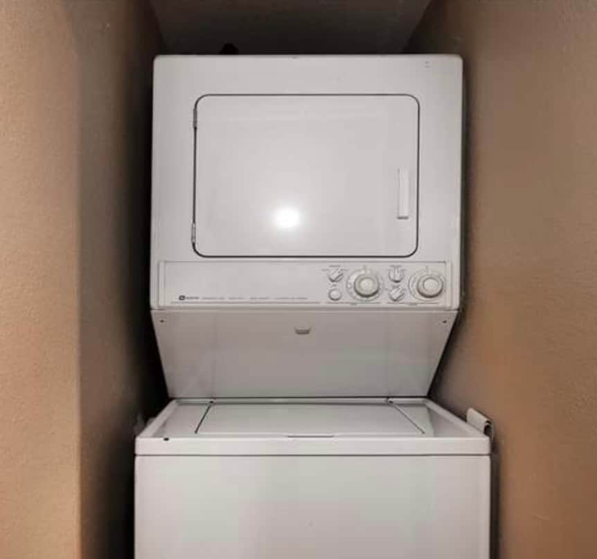A stacked washer and dryer unit is located in a compact laundry space. The machine features a white exterior with control knobs and a door for easy access. The walls are painted in warm tones, providing a simple yet functional area.