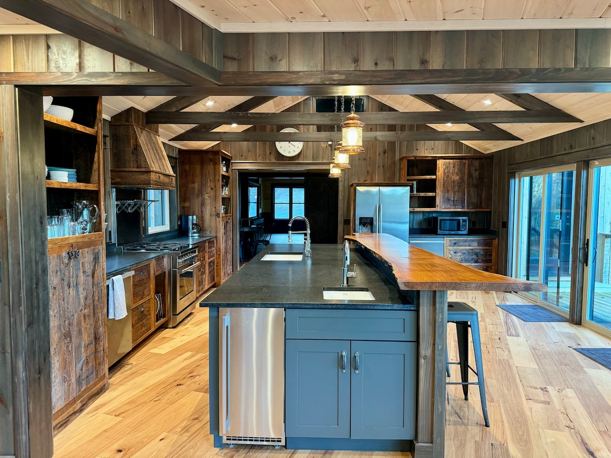 The spacious kitchen features wooden beam ceilings and rustic cabinetry, complemented by modern stainless steel appliances. A large central island houses a sink and has seating along one side. Sliding glass doors allow natural light and provide access to the outdoor deck.