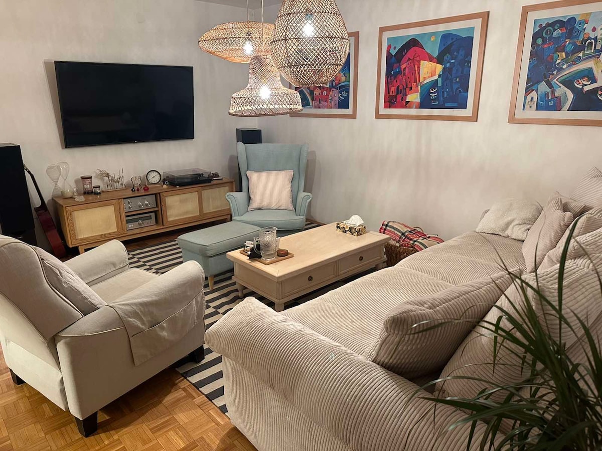 A cozy living area is presented with a spacious sectional sofa and an armchair in soft upholstery. A light-colored coffee table is centrally positioned, surrounded by decorative accents. Three woven pendant lights illuminate the space, which features vibrant artwork on the walls and a patterned rug.