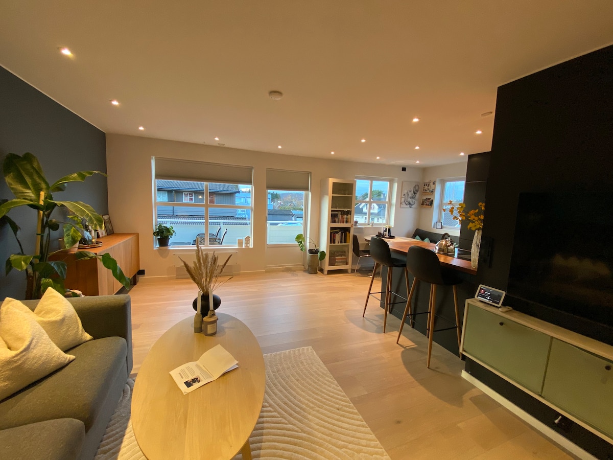 A modern living space is highlighted by a soft gray sofa and a round coffee table. Natural light flows in through large windows, illuminating the light wood flooring. A dining area with high chairs is visible, along with decorative plants and shelves showcasing books and decor.