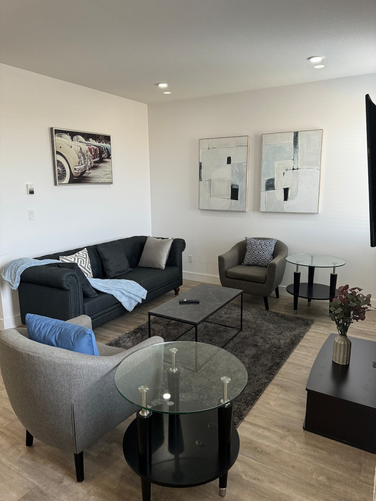 A modern living area features a sleek black sofa adorned with a soft throw blanket. Two accent chairs are positioned beside a glass coffee table atop a textured rug. Abstract artwork hangs on the walls, enhancing the bright and airy ambiance.