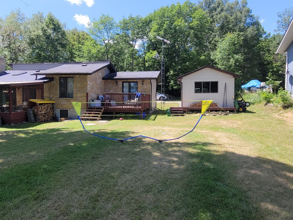 A spacious grassy yard is presented, bordered by trees, with a deck leading to a cottage and a storage shed in the background. Equipment for outdoor activities is visible. The area offers ample space for various recreational activities.