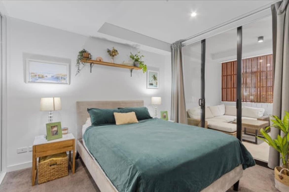 A spacious bedroom features a soft, green bedding set atop a comfortable bed. Two bedside lamps provide gentle lighting, while decorative plants and art enhance the decor. Glass doors lead to a separate lounge area, allowing natural light to fill the space.