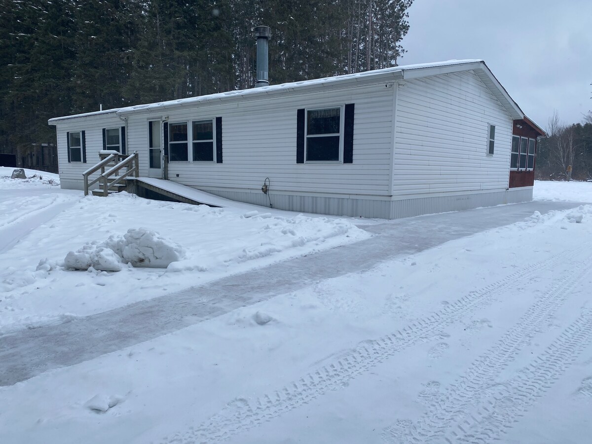 The exterior of a single-story home is shown, featuring a light-colored facade and black window shutters. Snow blankets the surrounding ground, while a wooden porch step leads to the entrance. A clear driveway extends towards the home, flanked by trees in the background.
