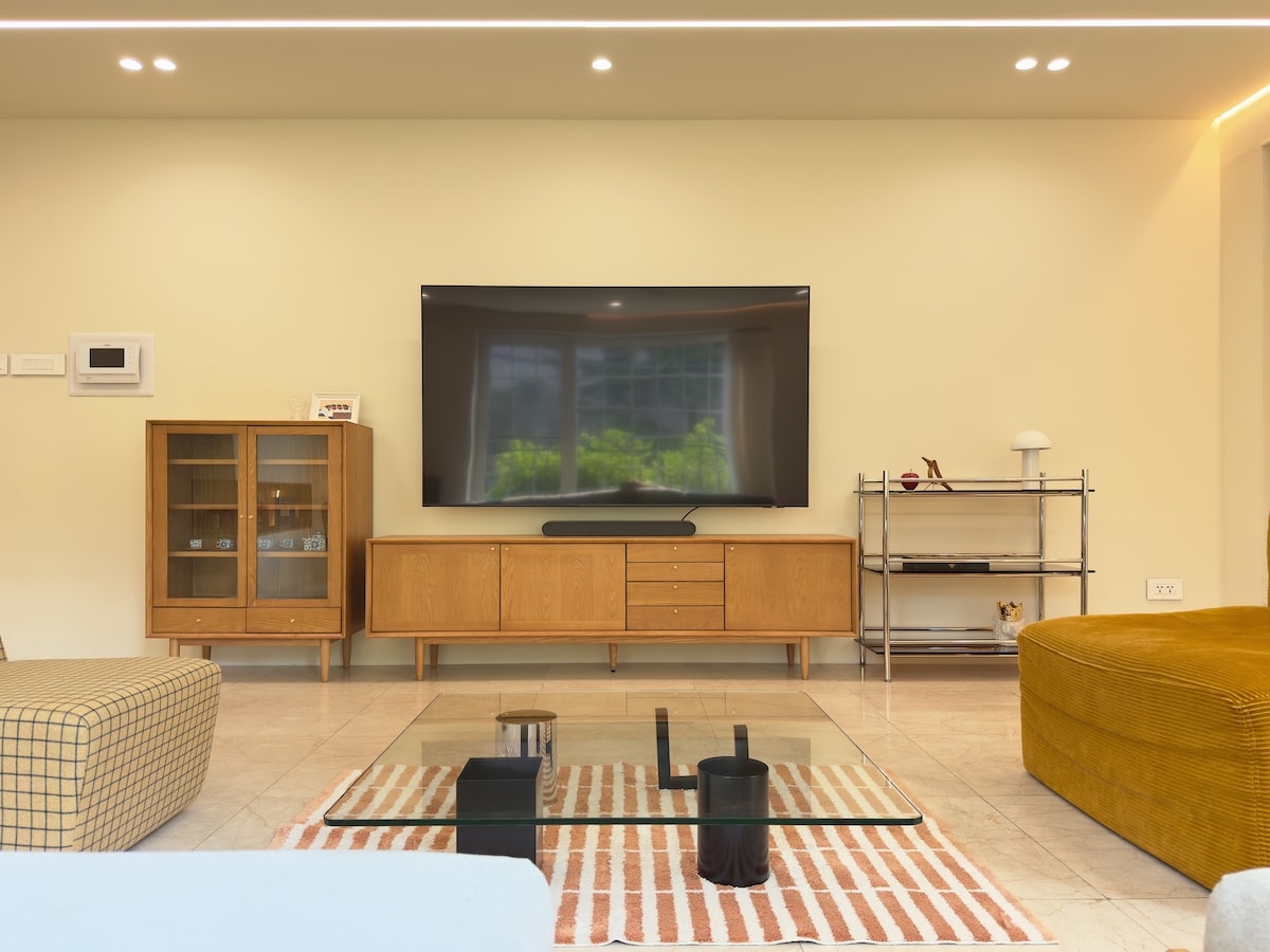 The living room features a modern design with a large, wall-mounted TV. A sleek glass coffee table is placed on a striped rug, surrounded by plush seating options in various shapes. Warm lighting enhances the spacious ambiance, while a cabinet and shelving unit provide functional decor.