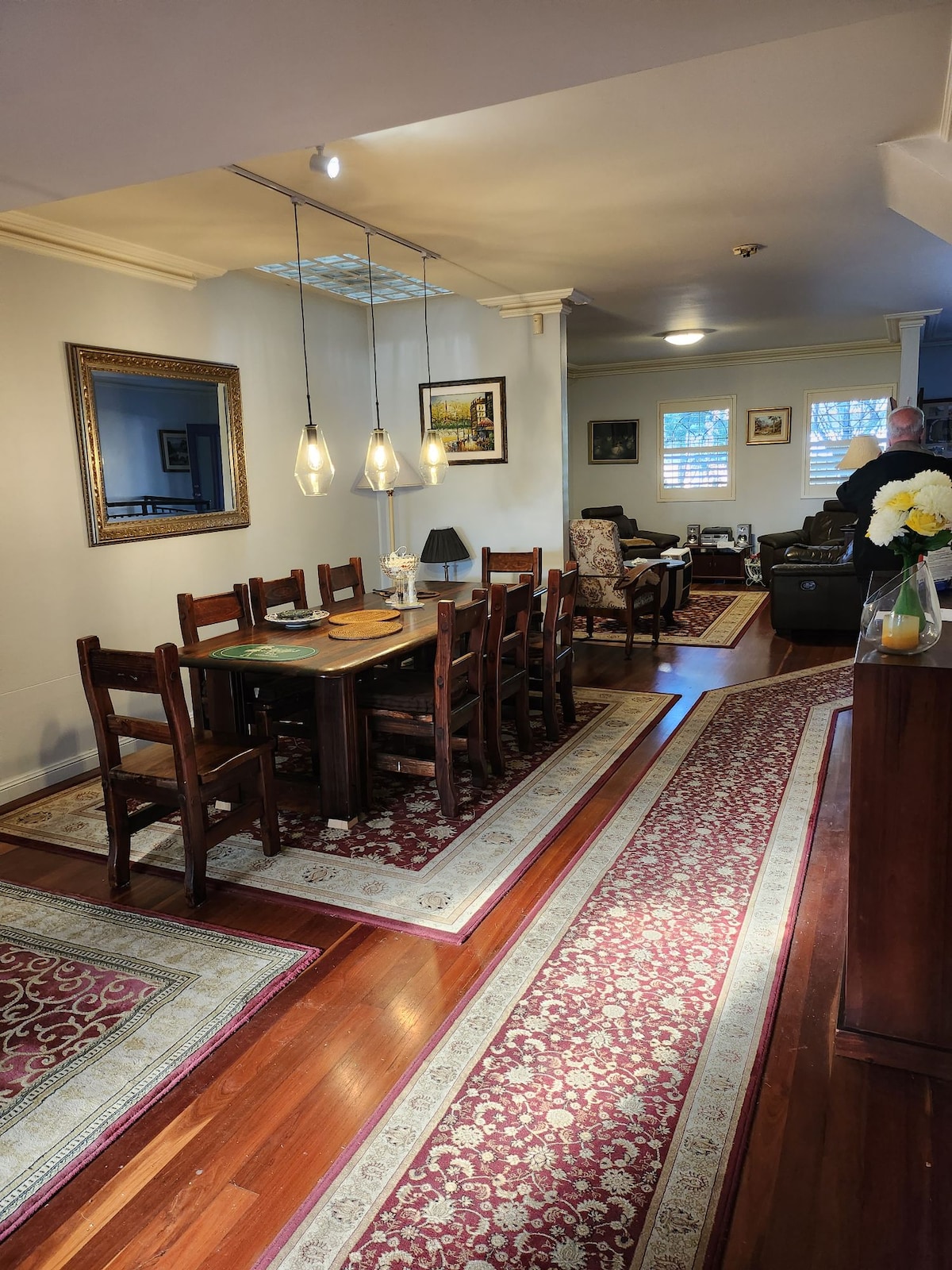 A dining area features a large wooden table surrounded by eight chairs, illuminated by three pendant lights above. The space is complemented by two area rugs and a decorative mirror on the wall, creating a welcoming environment for gatherings.