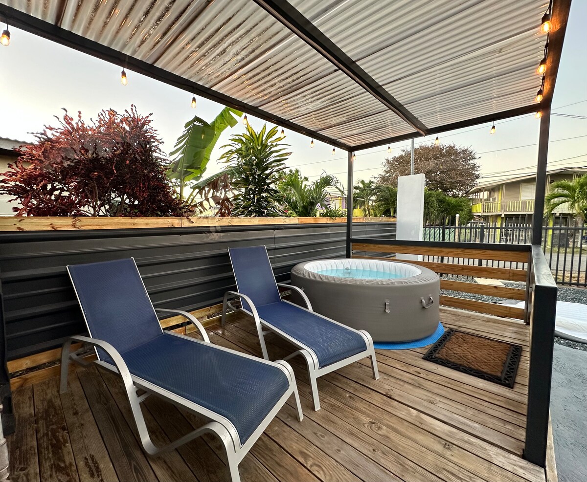 A shaded outdoor area features two lounge chairs alongside a circular hot tub. Greenery surrounds the space, with decorative plants enhancing the atmosphere. Soft lighting is provided by string lights overhead, creating a relaxing environment for enjoyment.