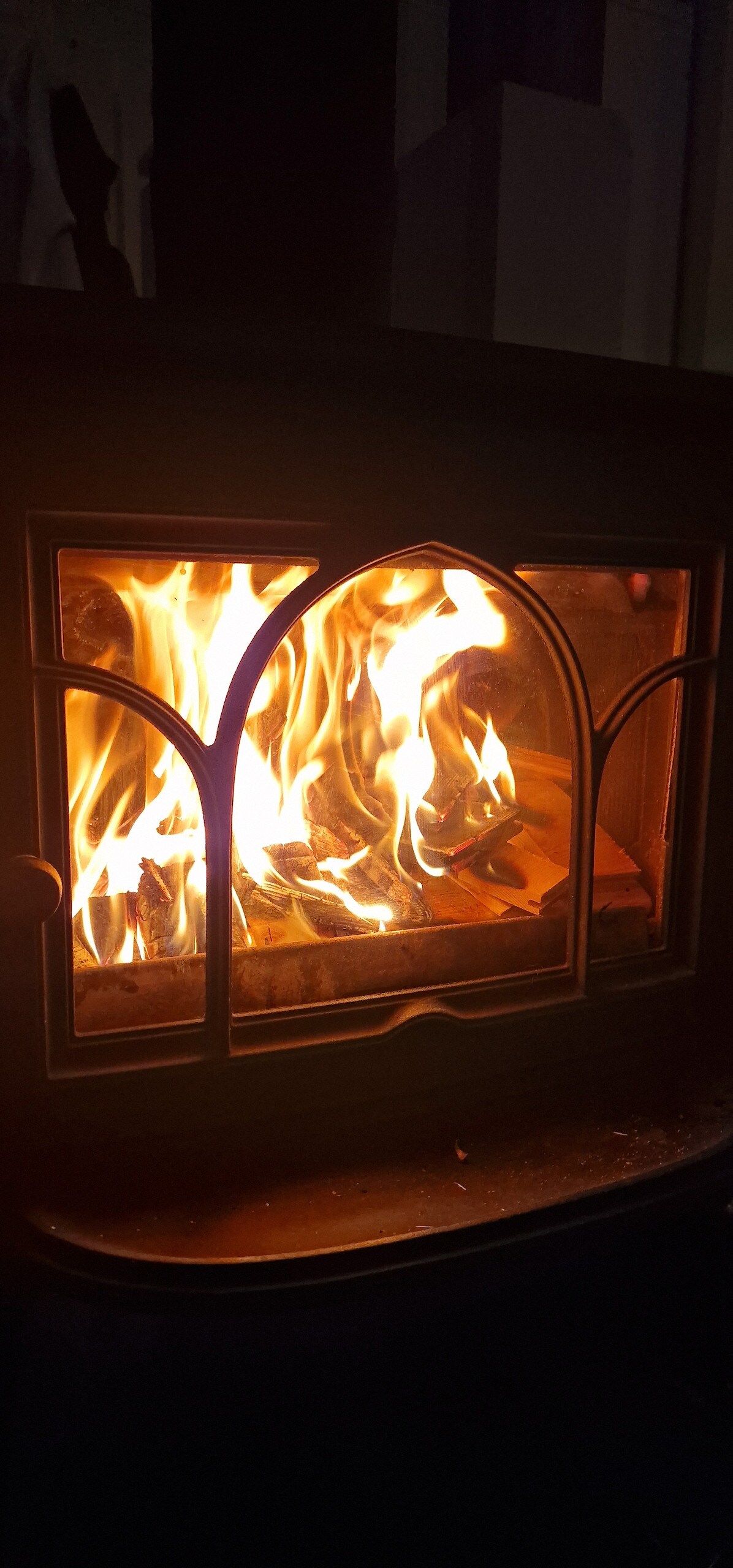 A warm glow is emitted from the fireplace, where flames dance and flicker inside a glass-fronted stove. The surrounding darkened space enhances the inviting ambiance created by the fire.