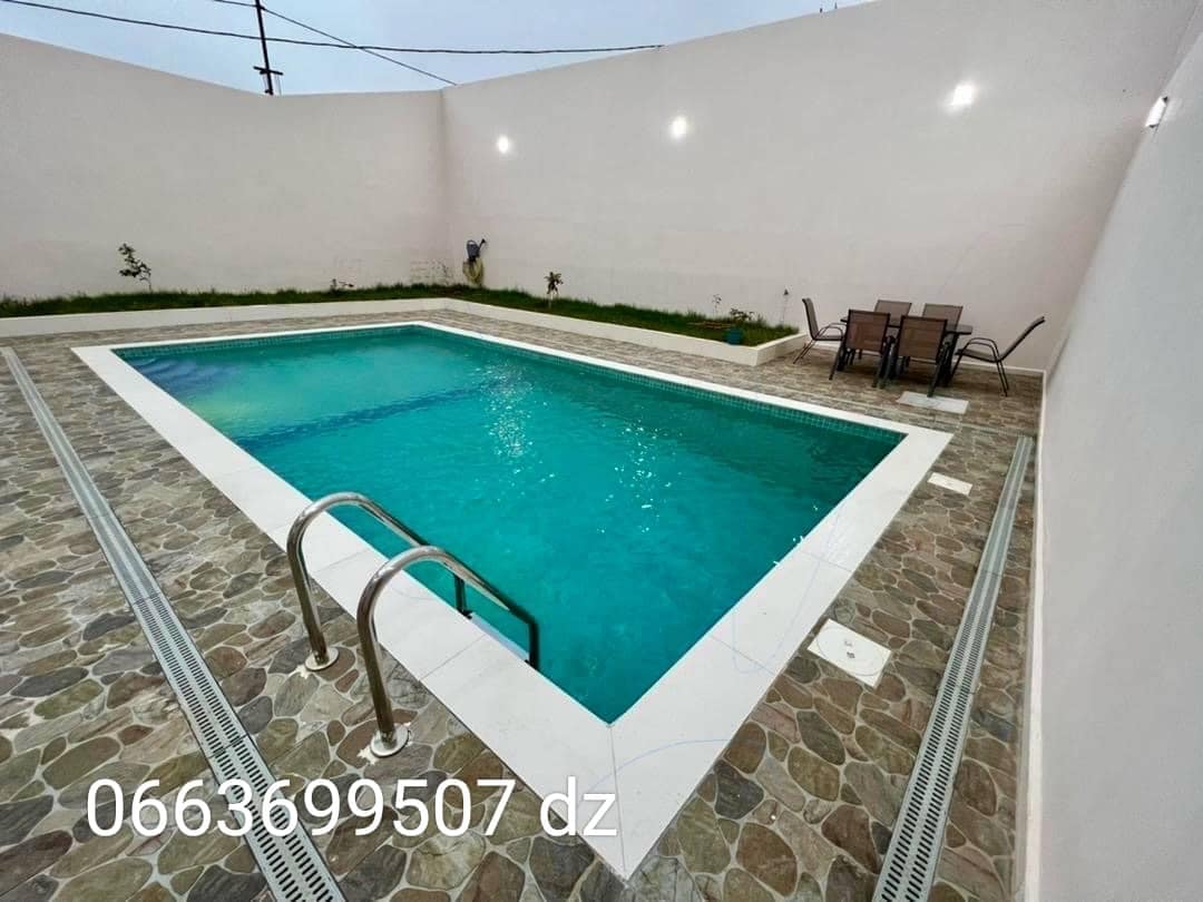 A rectangular swimming pool is surrounded by a stone patio, featuring steps leading into the water. Comfortable seating is arranged nearby, accommodating several guests. The pool is well-lit, enhancing its inviting appearance during twilight.
