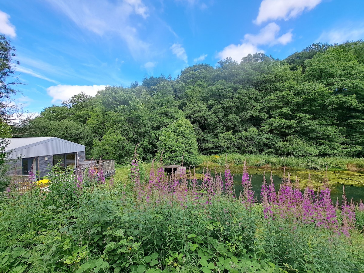 A tranquil scene captures the cabin nestled among vibrant greenery and wildflowers, with soft pink blossoms in the foreground. The serene lake reflects the surrounding lush trees under a bright blue sky, creating a peaceful retreat in nature.
