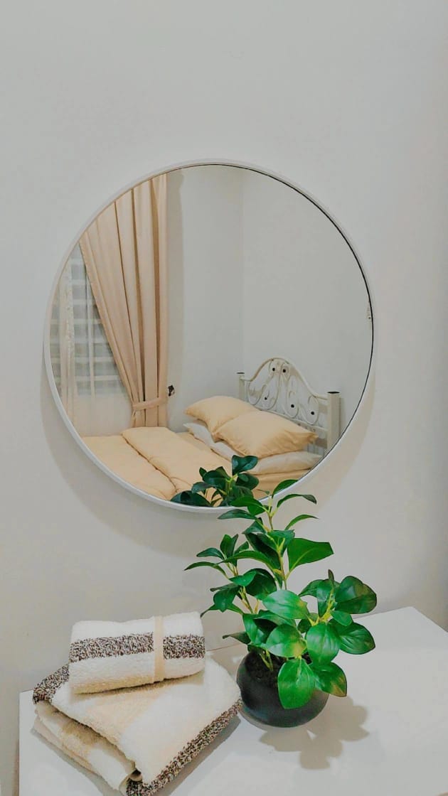 A round mirror reflects a neatly made bed with soft pillows and light-colored bedding. In front of the mirror, a small potted plant sits atop a table, accompanied by neatly folded towels in neutral tones, creating a tidy and appealing arrangement.