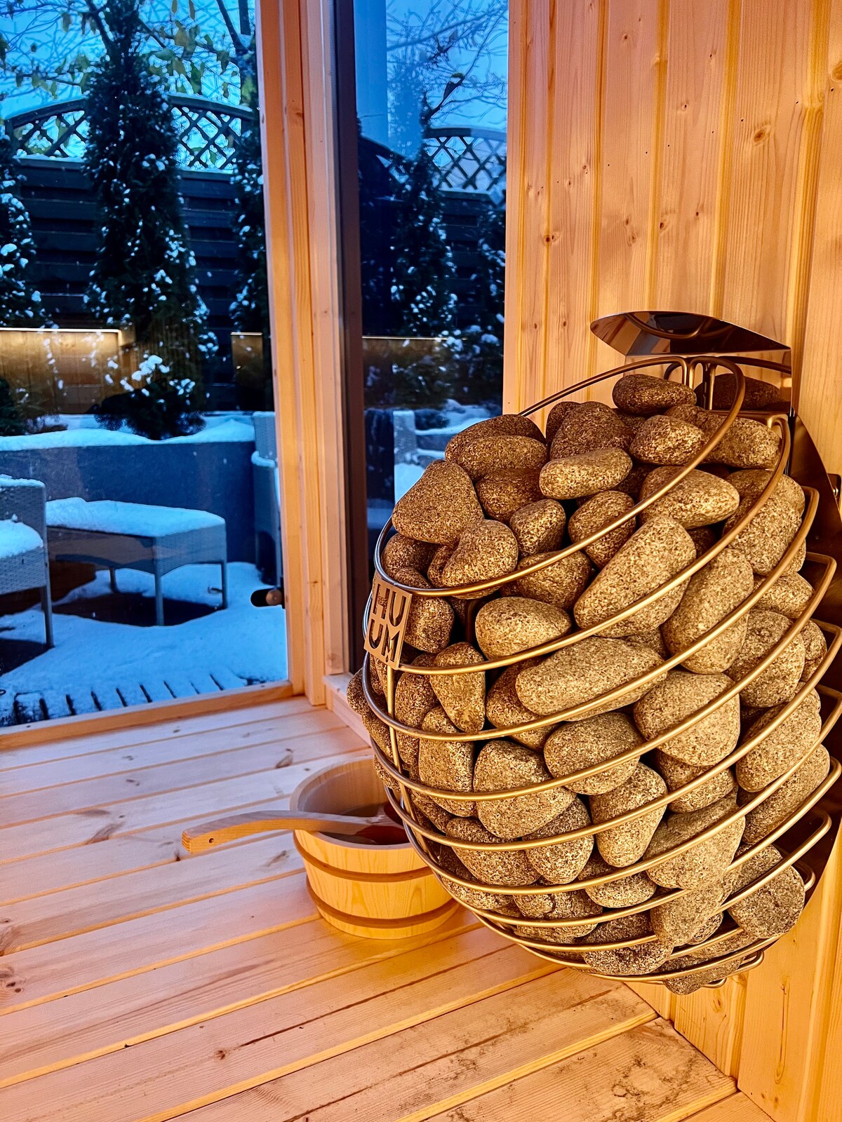 A wooden sauna interior features a circular arrangement of smooth stones for steam generation. A wooden bucket sits nearby, and large windows reveal a snowy outdoor scene with landscaped areas. The warm wood tones create a cozy and calming ambiance.