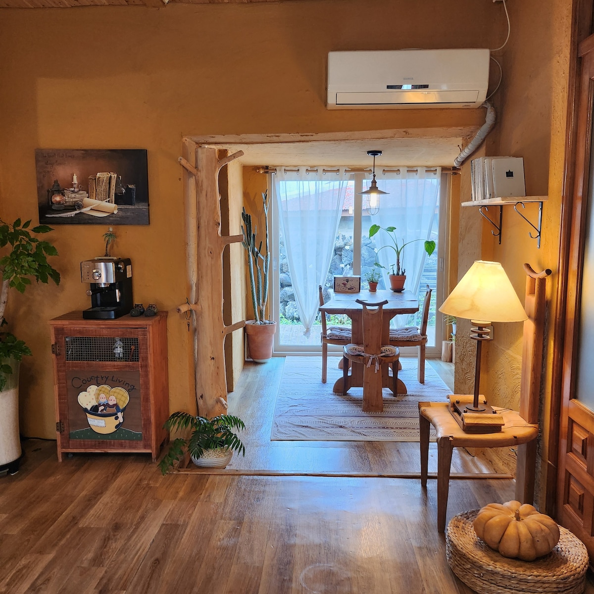 An inviting indoor space features earthy yellow walls made of natural materials, harmonizing with wooden furniture. A small dining table surrounded by chairs sits near a window adorned with sheer curtains, allowing soft natural light to filter through. A coffee maker and a bedside lamp add to the cozy ambiance.