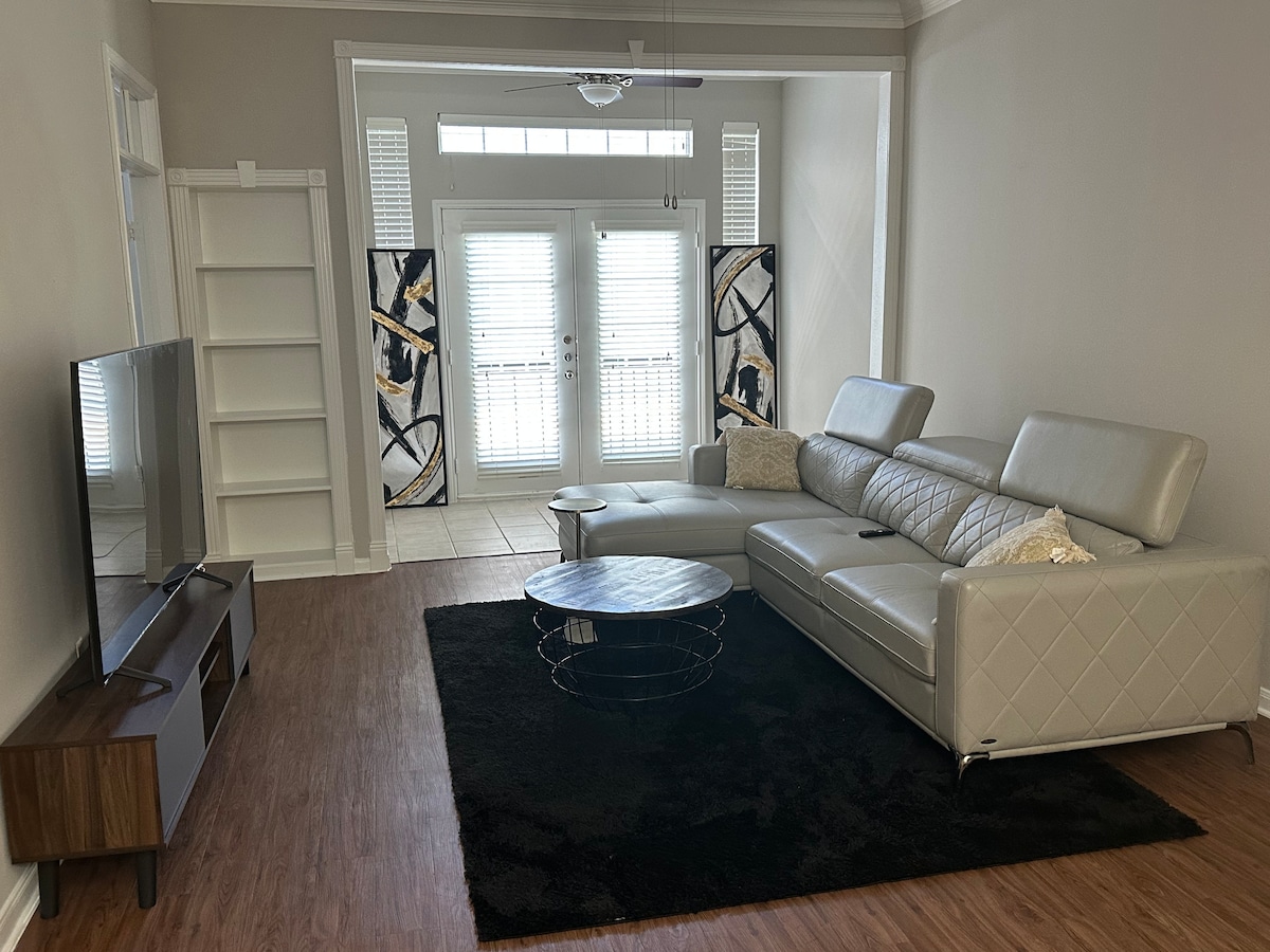 A modern living area features a spacious light gray sectional sofa positioned on a dark area rug. A sleek coffee table complements the setting, and a television is mounted on a wooden console against the wall. Natural light enters through several patio doors, which are adorned with blinds.