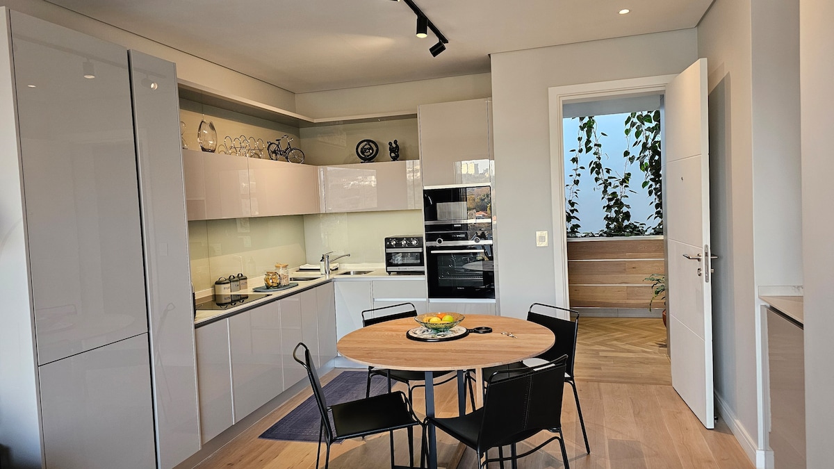 The modern kitchen is equipped with sleek cabinetry and stainless-steel appliances. A round dining table with four black chairs occupies the center, while natural light filters through a window adorned with indoor greenery. A neutral palette creates a clean and inviting space.