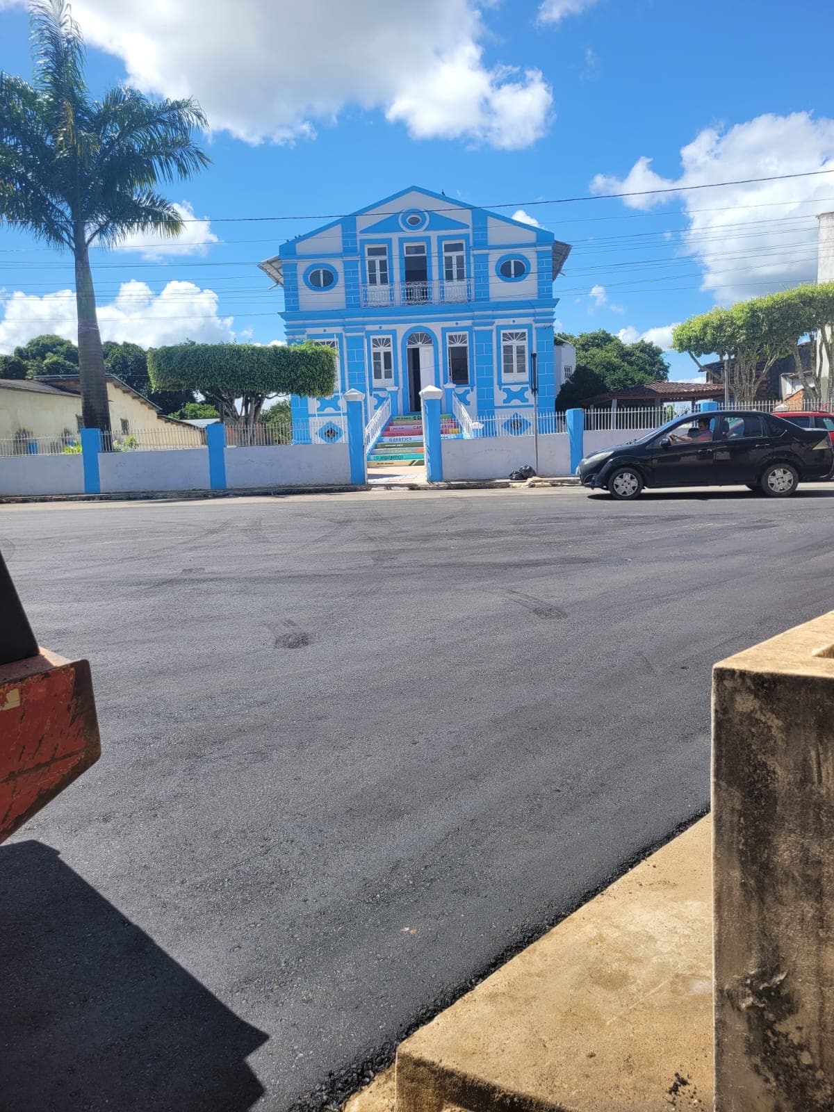 A vibrant blue house with intricate architectural details stands prominently against a sunny sky. Lush greenery and neatly trimmed hedges frame the property, while a well-maintained street provides easy access. The scene reflects a charming neighborhood setting with a welcoming atmosphere.