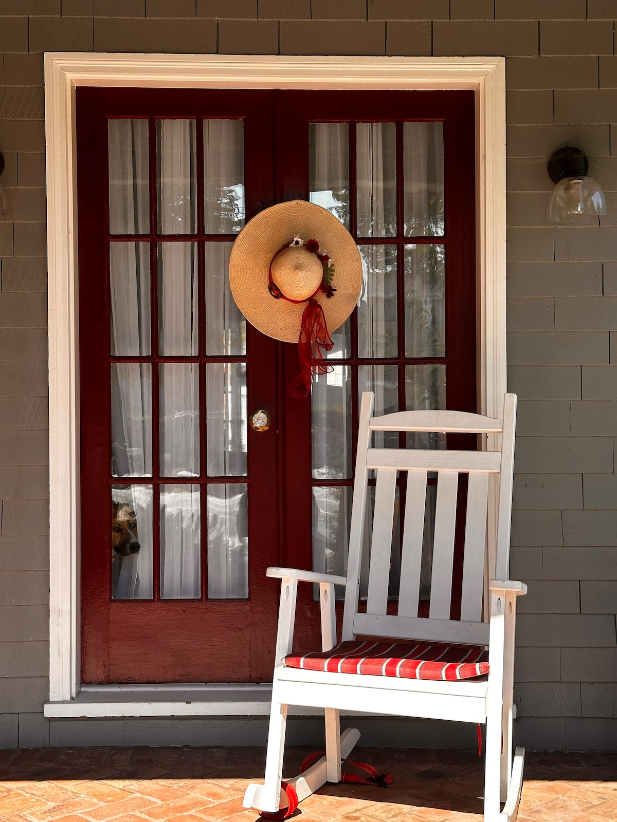 A charming entryway is showcased with red double doors featuring glass panels, adorned with a straw hat. A white rocking chair with red and white striped fabric is positioned nearby, inviting relaxation. Natural light enhances the warmth of the space.