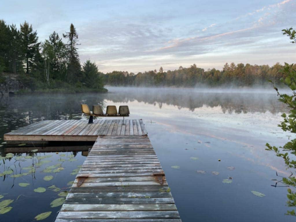 A wooden dock extends into a tranquil lake, surrounded by lush greenery and morning mist. Three chairs are positioned near the edge, overlooking the still waters, while lily pads float gently in the foreground, creating a peaceful natural setting.
