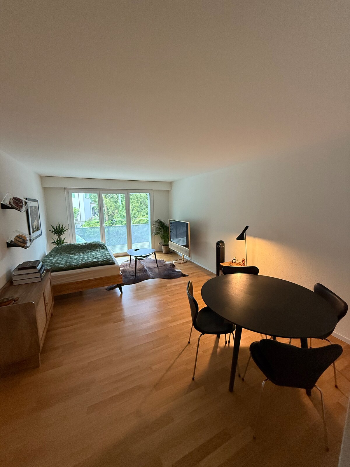 A spacious studio apartment interior is shown, featuring a deluxe sleeping sofa beside a wooden cabinet. A round black dining table and chairs are positioned nearby. Large windows allow natural light to fill the room, which is complemented by warm wood flooring.