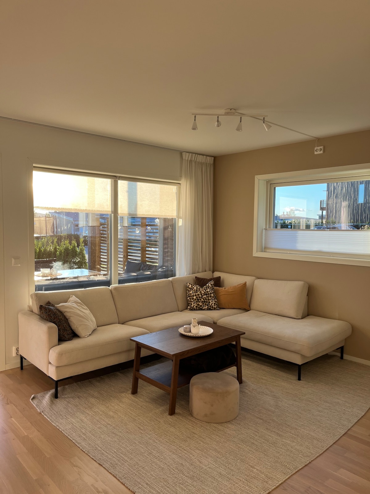 A light-filled living area features a neutral sectional sofa with accent pillows, oriented toward a wooden coffee table. Natural light pours in through large windows, offering views of the outdoor space. A subtle area rug adds warmth to the hardwood floor.