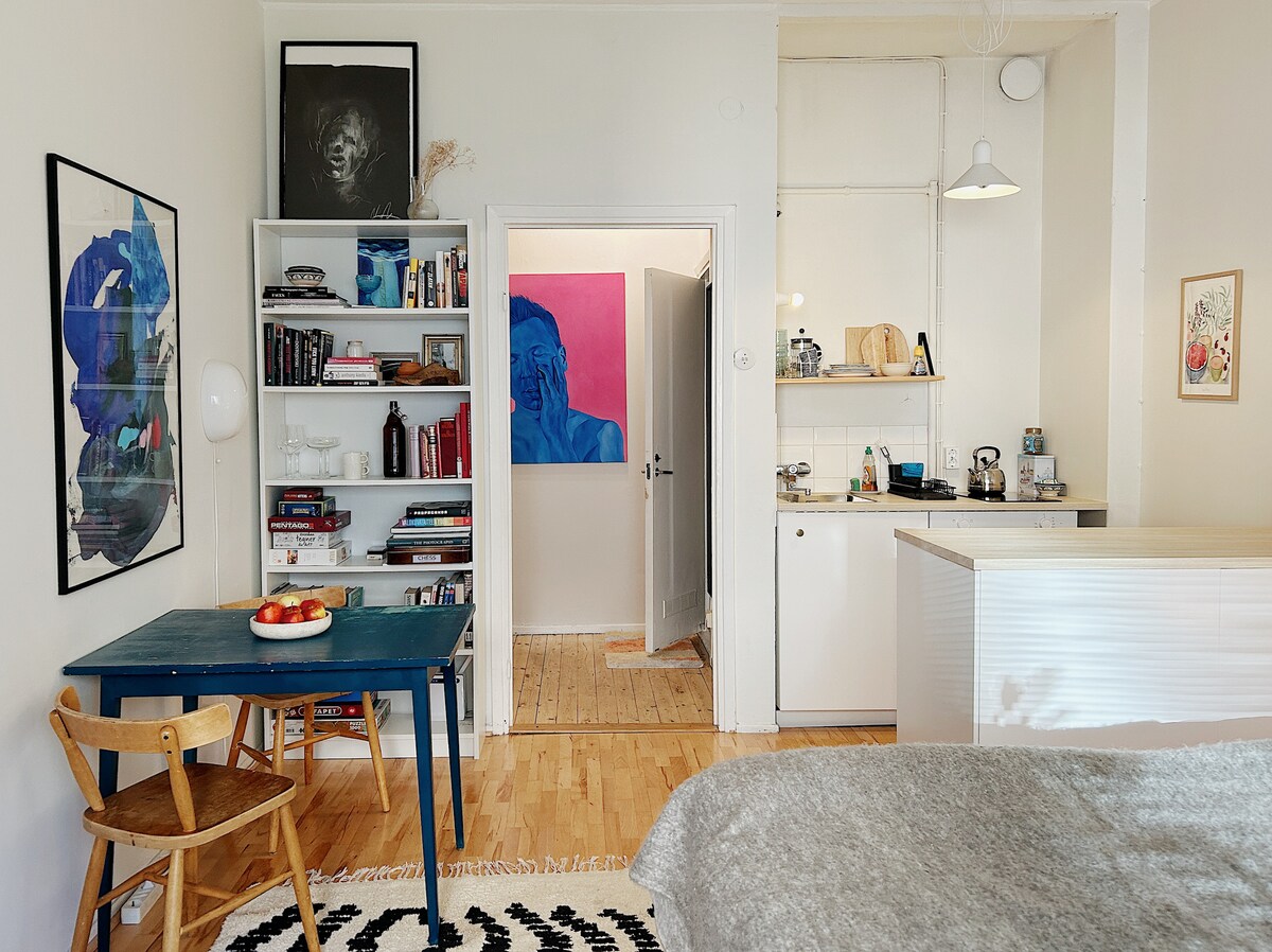 A cozy one-room apartment is displayed, featuring a blue table with wooden chairs and a small bookshelf filled with books. An open kitchen area is visible at the back, with modern appliances. The walls are adorned with colorful artwork, and a light oak floor enhances the space.