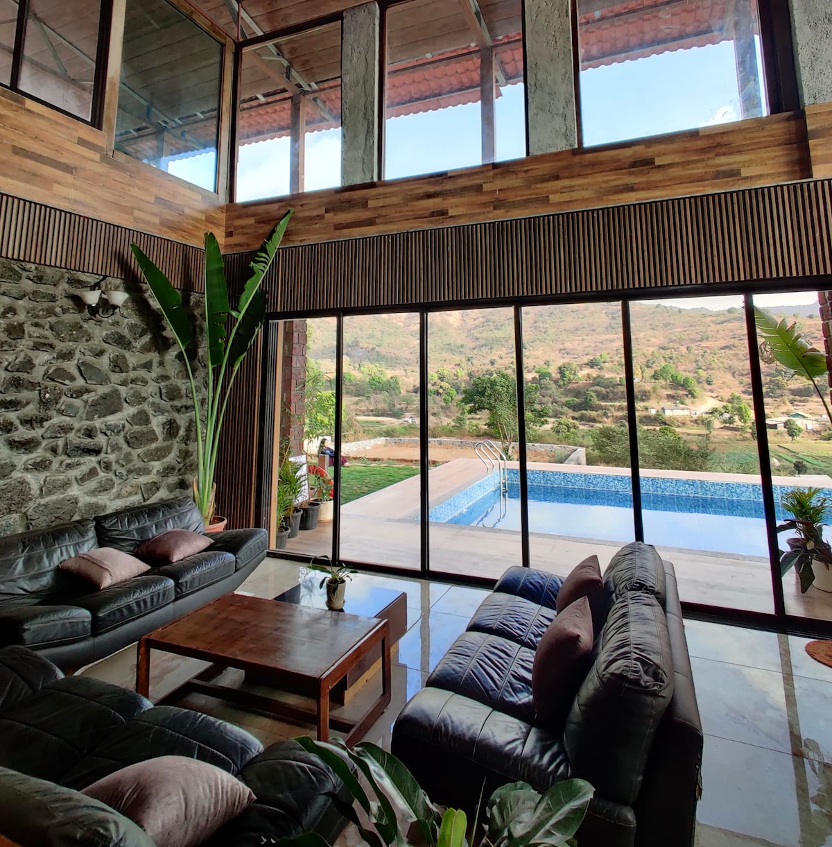 A spacious living area features large glass windows offering views of the surrounding mountains and a swimming pool. Comfortable seating arrangements are positioned beside a wooden coffee table, with indoor plants adding a touch of greenery to the inviting atmosphere.