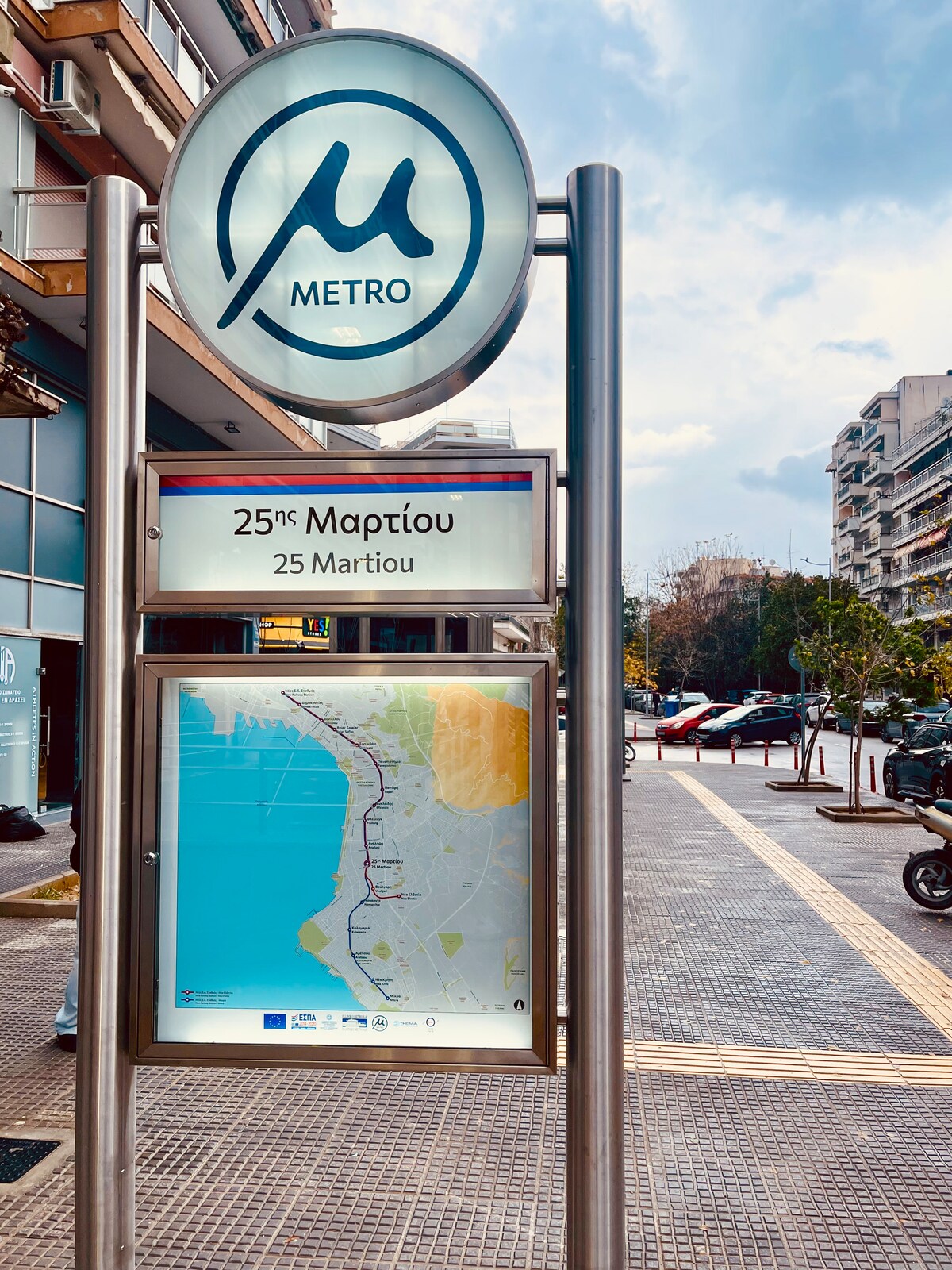 A metropolitan transit sign displaying '25 Μαρτίου' and a detailed map of the area is prominently featured. The surrounding streets are lined with trees and buildings, indicating a busy urban environment. Nearby, parked vehicles add to the lively atmosphere of the neighborhood.