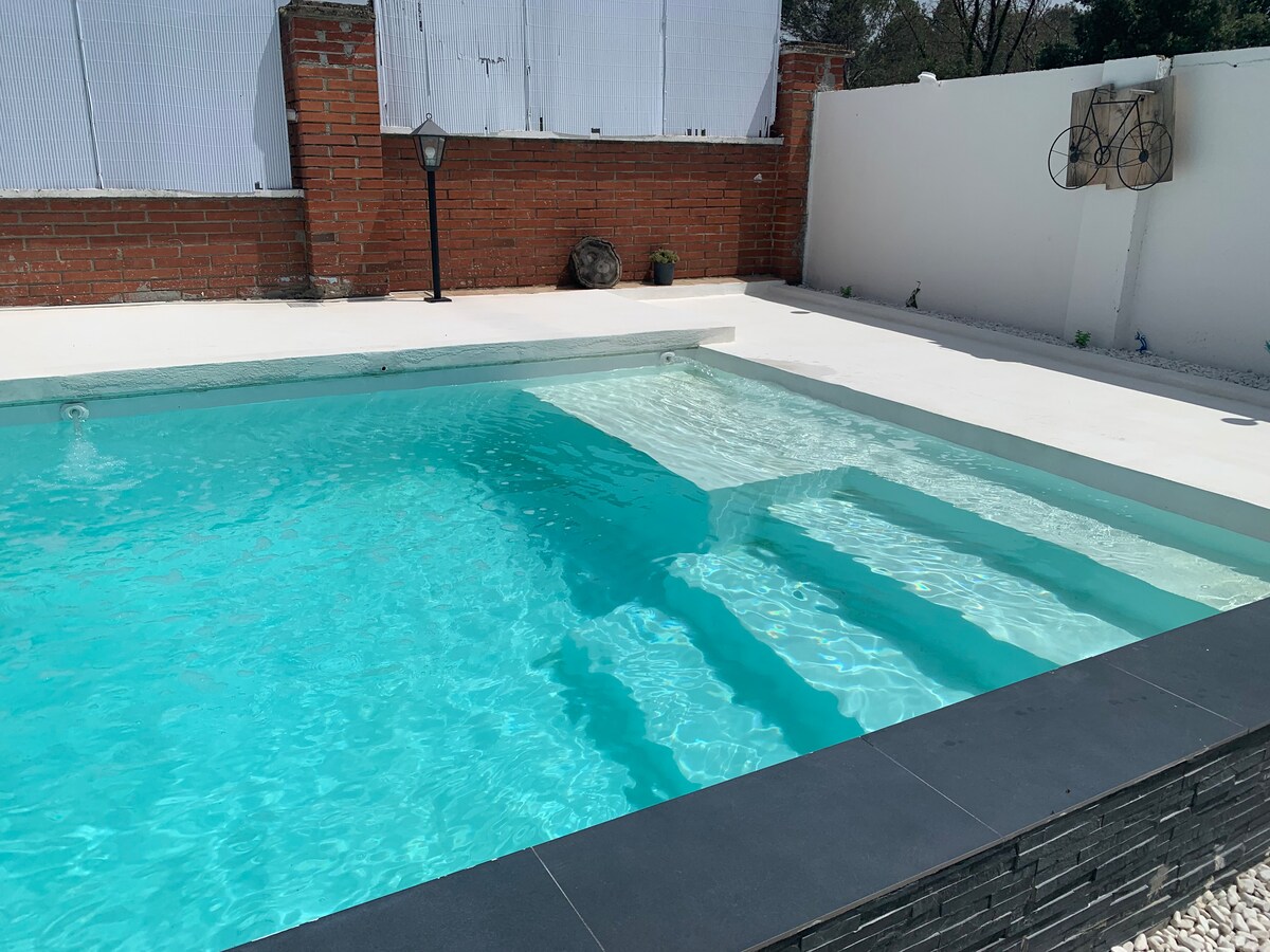 A shared swimming pool is visible, featuring clear turquoise water reflecting sunlight. The surrounding area includes a smooth white deck and a brick wall, enhancing the open space. A lamp post is positioned nearby, adding functionality to the inviting setting.