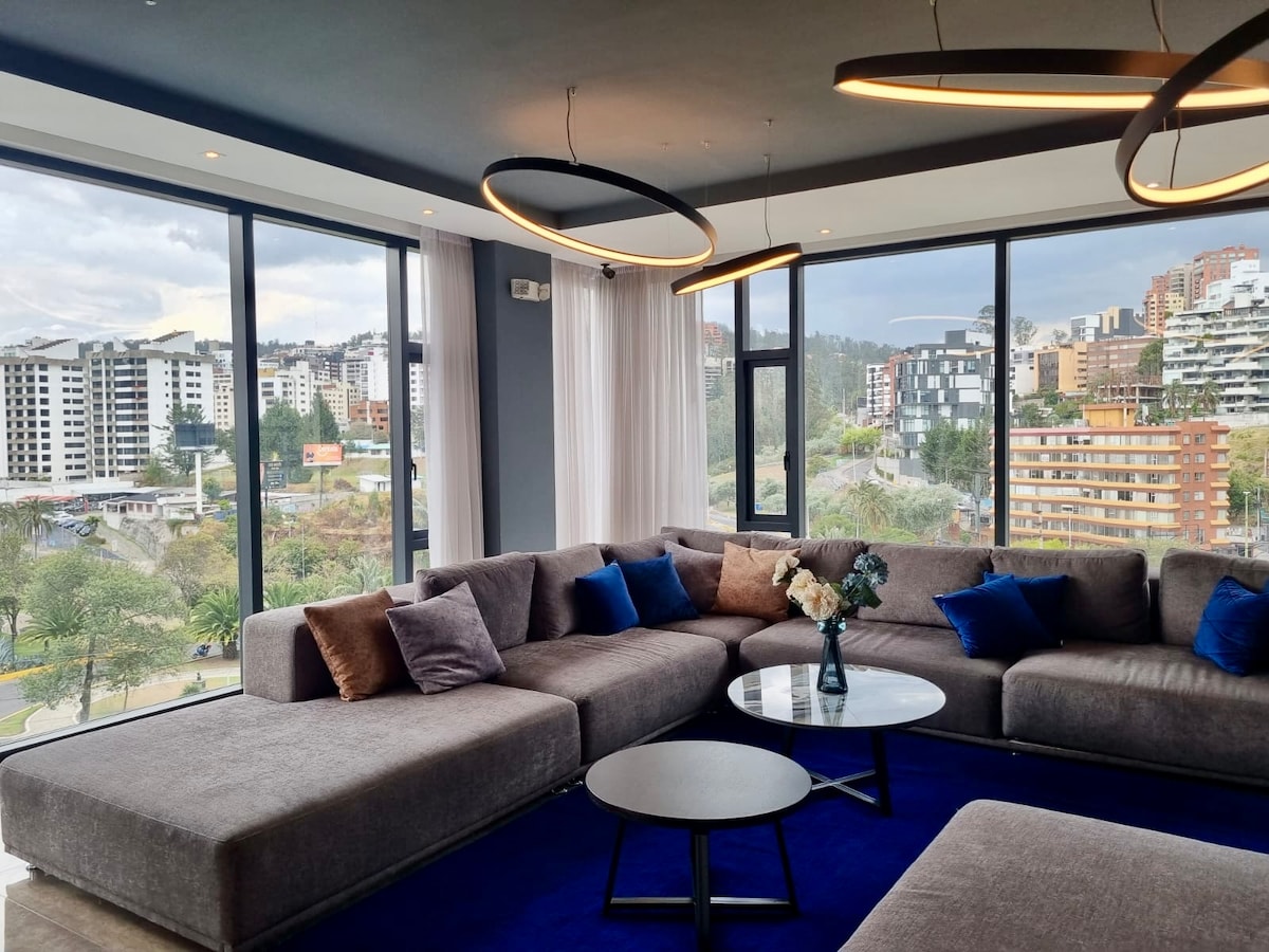 A spacious lounge area features large windows that frame a view of the city and surrounding greenery. A sectional sofa adorned with plush pillows offers comfortable seating. A coffee table sits centered on a deep blue area rug, enhancing the room's modern aesthetic.