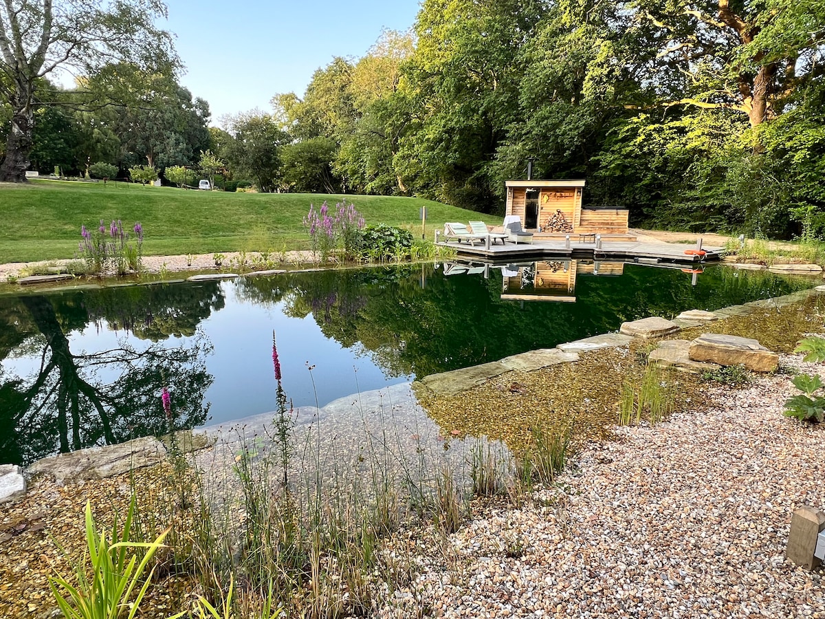 A crystal-clear swimming pond reflects the surrounding greenery, bordered by a pebbled shore and patches of aquatic plants. A wooden sauna building and deck area are positioned nearby, providing a serene space for relaxation amidst the natural setting.